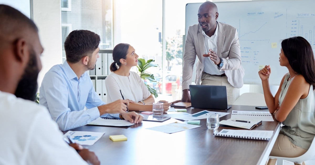 Featured image for "Digital Marketing for Staffing Agencies: What Top Agencies Are Doing for a Successful 2023" blog post. Four people are sitting around a table with paperwork in front of them in a conference room while one man in a suit is standing and speaking.