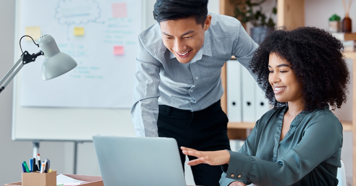 Featured image for "Using Your Staffing Website to Drive New Sales" blog post. Two coworkers are looking at a computer screen, the woman is sitting while the man is standing next to her.
